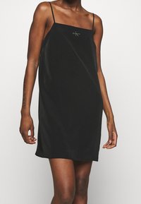 Schwarzes Satinkleid mit dünnen verstellbaren Trägern, geradem Schnitt und einem dezenten Logo-Detail auf der Vorderseite. Glattes, minimalistisches Design.