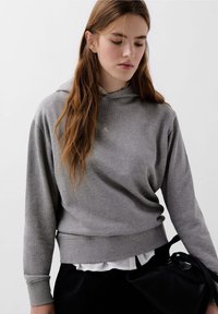 Felpa grigia in cotone con una vestibilità rilassata, dotata di cappuccio con coulisse e polsini a coste. Abbinata a una camicia bianca a strati sotto.