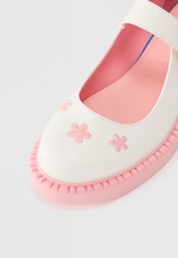 White leather shoe with pink floral embroidery, round toe, and pink rubber sole featuring a textured edge and strap across the instep.