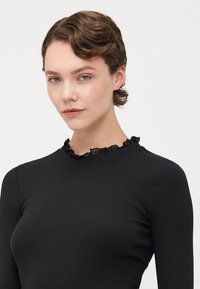 Black ribbed long-sleeve top with a textured ruffled neckline, fitted design, and smooth fabric, highlighting a minimalist aesthetic.