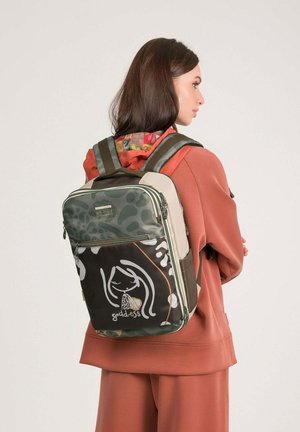 Femme portant un sweat à capuche et un pantalon couleur rouille, portant un sac à dos vert et beige avec le texte "goddess" et un visage illustré sur la poche avant.