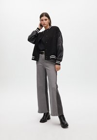 Černá bomber bunda s lesklými rukávy, bílými pruhy a písmenovým detailem. Kombinována s vysoce střiženými šedými širokými džíny a černými botami.