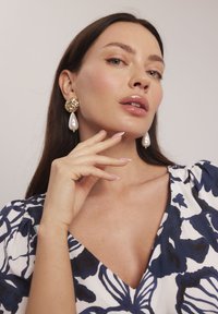 Złote kolczyki w wyrazistym stylu z fakturowanym okrągłym wierzchem i białą perłą na końcu. Modelka z długimi włosami i naturalnym makijażem pozuje z ręką przy twarzy.
