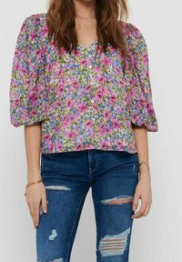 Blumenbluse in Lila und Grün, mit Puffärmeln, V-Ausschnitt und Knopfverschluss. Kombiniert mit dunkelblauen, ausgebleichten Jeans.