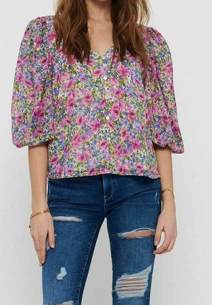 Blusa floreale in viola e verde, con maniche a sbuffo, scollatura a V e chiusura con bottoni. Abbinata a jeans scuri strappati.
