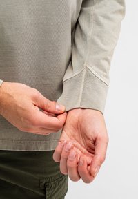 Helle beige Langarmshirt mit geripptem Bündchen und sichtbaren Nähten. Hände passen das Bündchen an und zeigen den weichen Stoff und den neutralen Farbton.