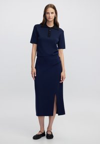 Žena stojící v námořnické modré krátkém rukávu s černým límečkem a sladěné dlouhé sukni s předním rozparkem, doplněné černými plochými botami.