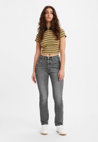 Gestreepte gele en marineblauwe cropped polo-tee, gecombineerd met hooggeritste grijze jeans. Model draagt witte sneakers en staat tegen een witte achtergrond.