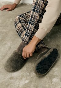 Ciemnozielone kapcie z futrem, o gładkiej powierzchni i wzorzystej czarnej gumowej podeszwie. Noszone z spodniami w kratę i strukturalnymi skarpetkami.