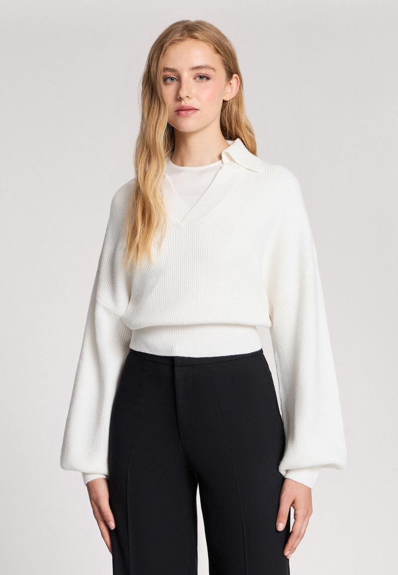 Giovane donna con lunghi capelli biondi che indossa un maglione bianco con scollo a V sopra una canottiera bianca e pantaloni neri su uno sfondo chiaro uniforme.