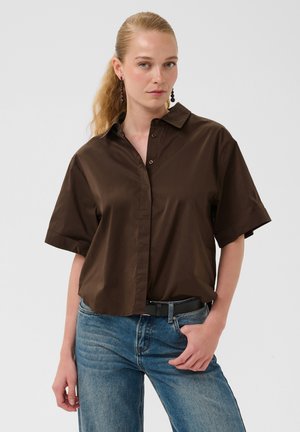 Femme à la peau claire et aux cheveux blonds attachés porte une chemise boutonnée manches courtes marron foncé et un jean bleu, debout avec une main dans la poche.