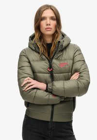 Giacca puffer verde oliva con cappuccio, dotata di zip frontale, polsini elasticizzati e un logo rosa sul lato sinistro del petto. Texture liscia e lucida.