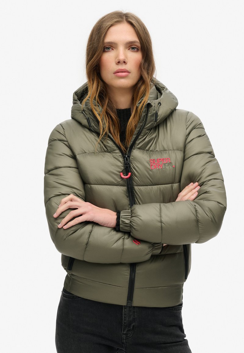 Olijfgroene pufferjack met een capuchon, voorzien van een ritssluiting aan de voorkant, elastische manchetten en een roze logo op de linkerkant van de borst. Smooth, glanzende textuur.
