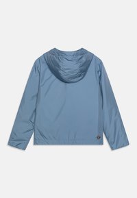 Giacca leggera blu con cappuccio, texture liscia, orlo elastico e un logo rotondo sul lato sinistro in basso.