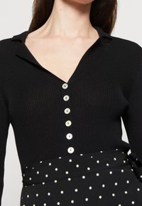 Černý žebrovaný cardigan s výstřihem do V a sedmi knoflíky v barvě perel po celé přední straně. Má dlouhé rukávy a přiléhavý střih, doplněný sukní s puntíky.