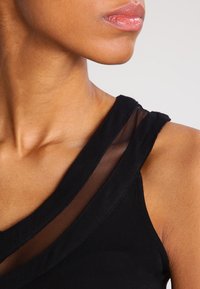Débardeur noir avec un design sans manches et un overlay en maille transparente au niveau de l'encolure. Texture lisse. Vue rapprochée de la zone de l'épaule.