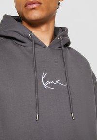 Grå hettegenser laget av mykt stoff, med en frontlomme, snorhette og hvit brodert logo på brystet.