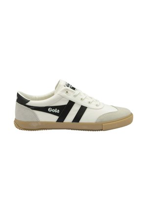 Zapatilla de lona blanca con rayas laterales y logotipo en negro, puntera de gamuza gris y suela de goma. Presenta seis ojetes con cordones blancos.