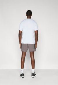 Λευκό κοντομάνικο t-shirt συνδυασμένο με γκρι σορτς που έχουν μαύρες πλευρικές ρίγες. Το μοντέλο είναι γυρισμένο με την πλάτη, φοράει μαύρα αθλητικά παπούτσια και λευκές κάλτσες.