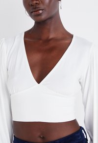 Biely crop top s hlbokým výstrihom do V a dlhými puff rukávmi, vyrobený z hladkej látky. Ševová časť pod prsiami v kombinácii s tmavými nohavicami.