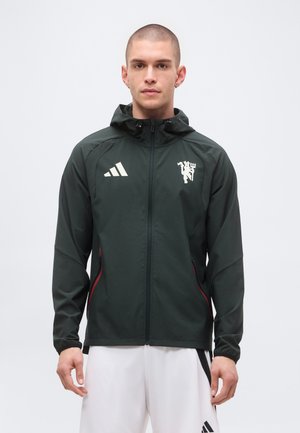 Giovane uomo che indossa una giacca nera con cappuccio e zip con loghi bianchi e pantaloncini sportivi bianchi su uno sfondo semplice.