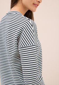 Eine Frau trägt einen strukturierten Pullover mit schwarz-weißen Streifen, zeigt ihre Schulter und das Profil von der Seite vor einem schlichten beigen Hintergrund.