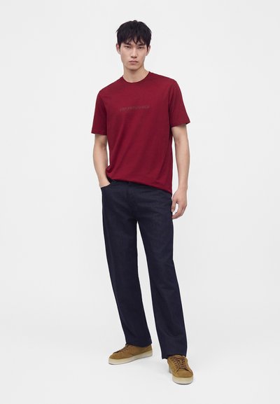 Joven de pie con una mano en el bolsillo, lleva una camisa roja de manga corta, pantalones oscuros y zapatillas marrones sobre un fondo blanco liso.