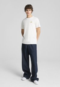 Camiseta de algodón blanca de manga corta, con un pequeño logo colorido en el pecho, combinada con unos vaqueros oscuros de denim holgados y zapatillas blancas.