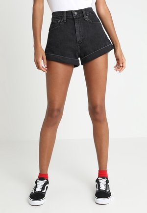 Femme portant un short en denim noir taille haute avec des ourlets retroussés, associé à des chaussettes rouges et des baskets noir et blanc, se tenant contre un fond blanc.