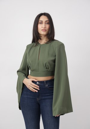 Femme aux longs cheveux foncés portant un haut court vert olive à manches larges et un jean bleu foncé, debout devant un fond clair uni.