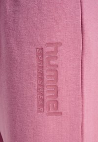 Lyserød sportstøjsstof med en tekstureret logo, der står "hummel" og "SPORTSWEAR" prentet på siden, som fremhæver den bløde tekstur.