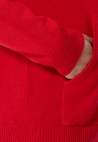 Roter Strickpullover mit gerippten Bündchen und einer vorderen Tasche. Weicher Stoff mit glatter Oberfläche, der klare Linien und ein lässiges Design betont.