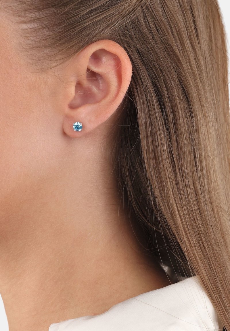 Gros plan sur l’oreille d’une personne aux cheveux bruns et raides, portant une petite boucle d’oreille clou ronde en pierre précieuse bleue.