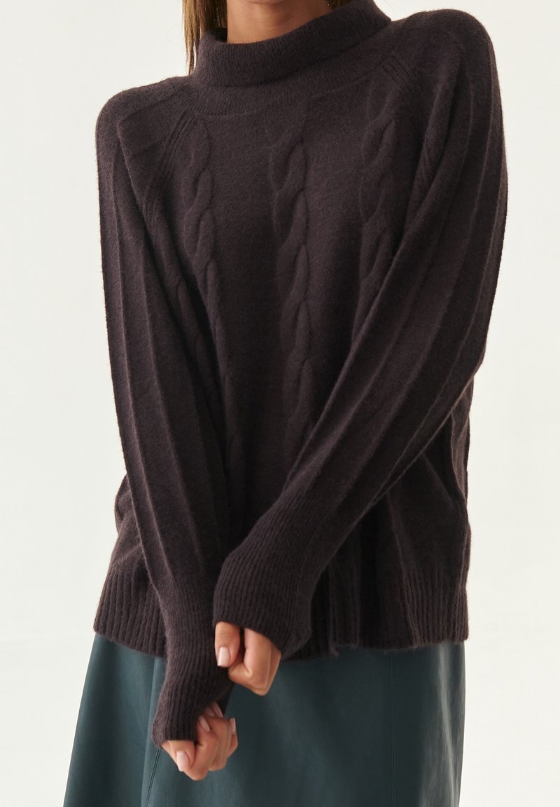 Pull en maille torsadée violet foncé avec un col haut, des poignets côtelés et une coupe décontractée, associé à une jupe verte. Texture douce évidente.