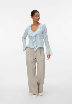 Blouse bleu clair à manches longues avec un col à volants et un motif floral, associée à un pantalon beige taille haute et des chaussures blanches.