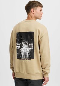 Beige sweatshirt met een zwarte grafische afbeelding van een stedelijke straatscene op de achterkant, met de tekst "BLEND SINCE 1993" eronder.