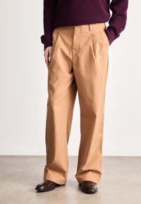 Beige, pamutkeverékből készült, pliszírozott nadrág, laza szabással. Sötétlila kötött pulóverrel és barna elegáns cipővel viselve.