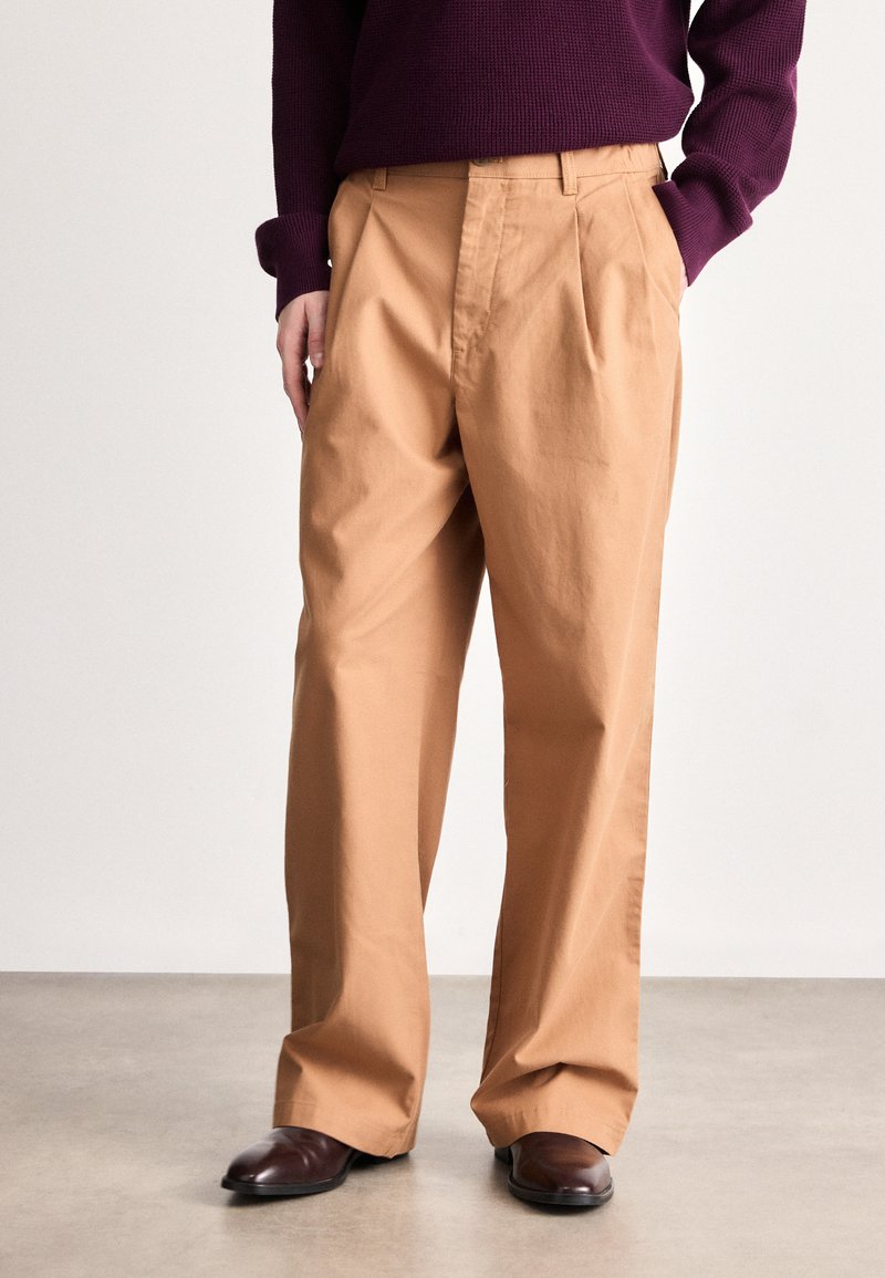 Beige, pamutkeverékből készült, pliszírozott nadrág, laza szabással. Sötétlila kötött pulóverrel és barna elegáns cipővel viselve.