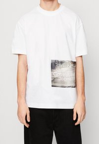 Bílé bavlněné tričko s černou obdélníkovou grafikou s textem „Calvin Klein Jeans“ v texturovaném potisku. Krátké rukávy a kulatý výstřih.