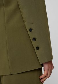 Õlivirooni värvi kohandatud jakk lähivaatlusel, millel on sile tekstuur, neli musta nuppu mansetil ja kontrastne tasku.