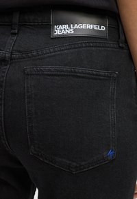Juodi džinsai išsiskiria ryškiu užpakaliniu kišeniu ir odinę etikete su baltu tekstu, kuris sako "KARL LAGERFELD JEANS." Matoma mėlyna siuvimo detalė.