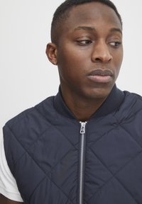 Marinblå, vadderad ärmlös jacka med ribbat krage och silverdragkedja. Buren över en vit t-shirt, med slät textur och diamantt mönster.