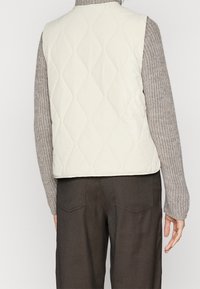 Prošívaná krémová vesta s hladkým povrchem. Má prošití ve vzoru diamantů, kulatý výstřih a volný střih, nošená přes pletený svetr.