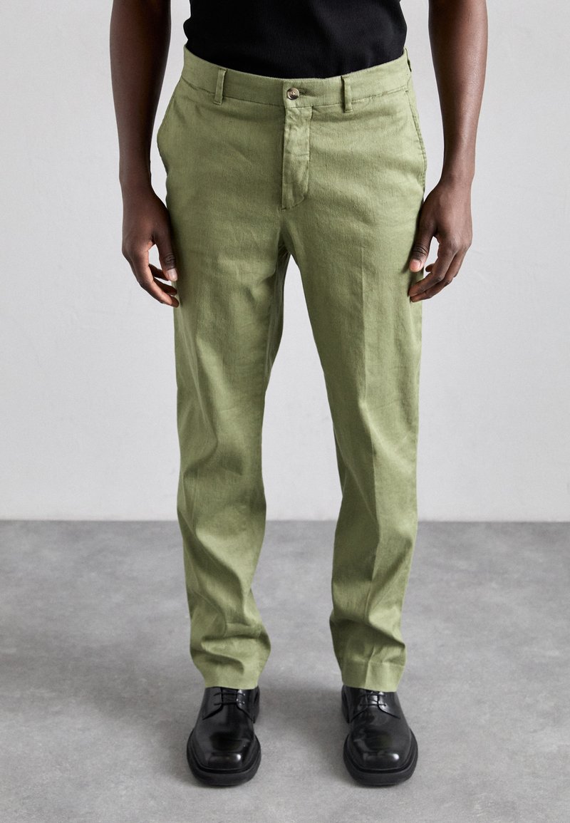 Groene linnen broek met een rechte snit, voorzien van een sluiting met een knoop aan de voorkant en zijzakken. Gecombineerd met zwarte leren schoenen.