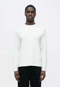 Baltas ilgomis rankovėmis marškinėlis su apvalia iškirpte, ant priekinės pusės subtiliai įspaustais užrašais. Lygus audinys, derinamas su tamsiomis kelnėmis.