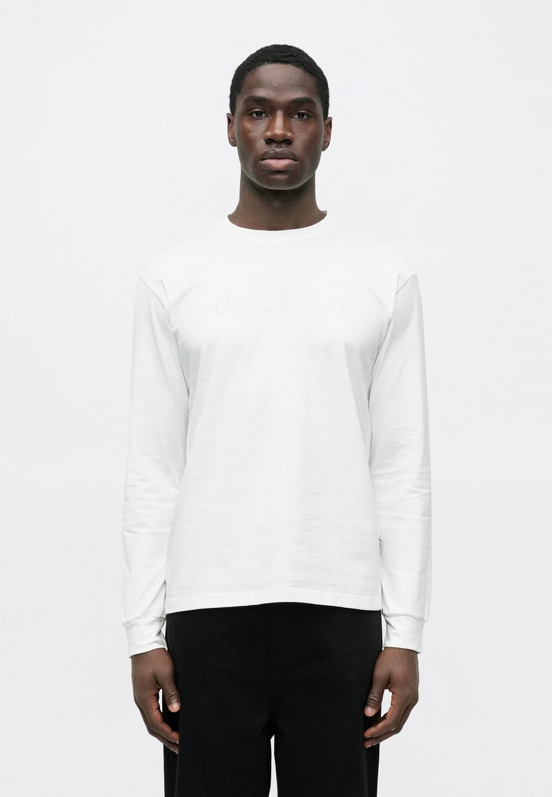 Baltas ilgomis rankovėmis marškinėlis su apvalia iškirpte, ant priekinės pusės subtiliai įspaustais užrašais. Lygus audinys, derinamas su tamsiomis kelnėmis.