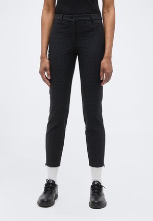 Kobieta w dopasowanych czarnych spodniach z subtelnym wzorem, białych skarpetkach i czarnych sznurowanych butach, stojąca na tle jasnego, jednolitego tła.