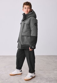 Halli veekindel jope mustade detailide, suurte esitaskute ja kapuutsiga. Koos mustade taskupükste ja valgete kingadega, millel on kummist tallad.