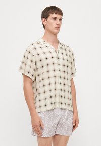 Camicetta a maniche corte, con bottoni, di colore beige chiaro con un motivo a griglia nera; tessuto leggero e trasparente, vestibilità comoda e design con colletto aperto.