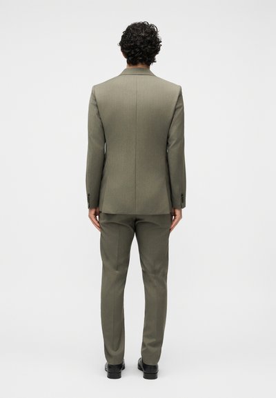 Άνδρας με σγουρά μαλλιά, φορώντας εφαρμοστό ελαφρώς πράσινο κοστούμι και μαύρα παπούτσια, στέκεται γυρισμένος με την πλάτη σε απλό φωτεινό φόντο.